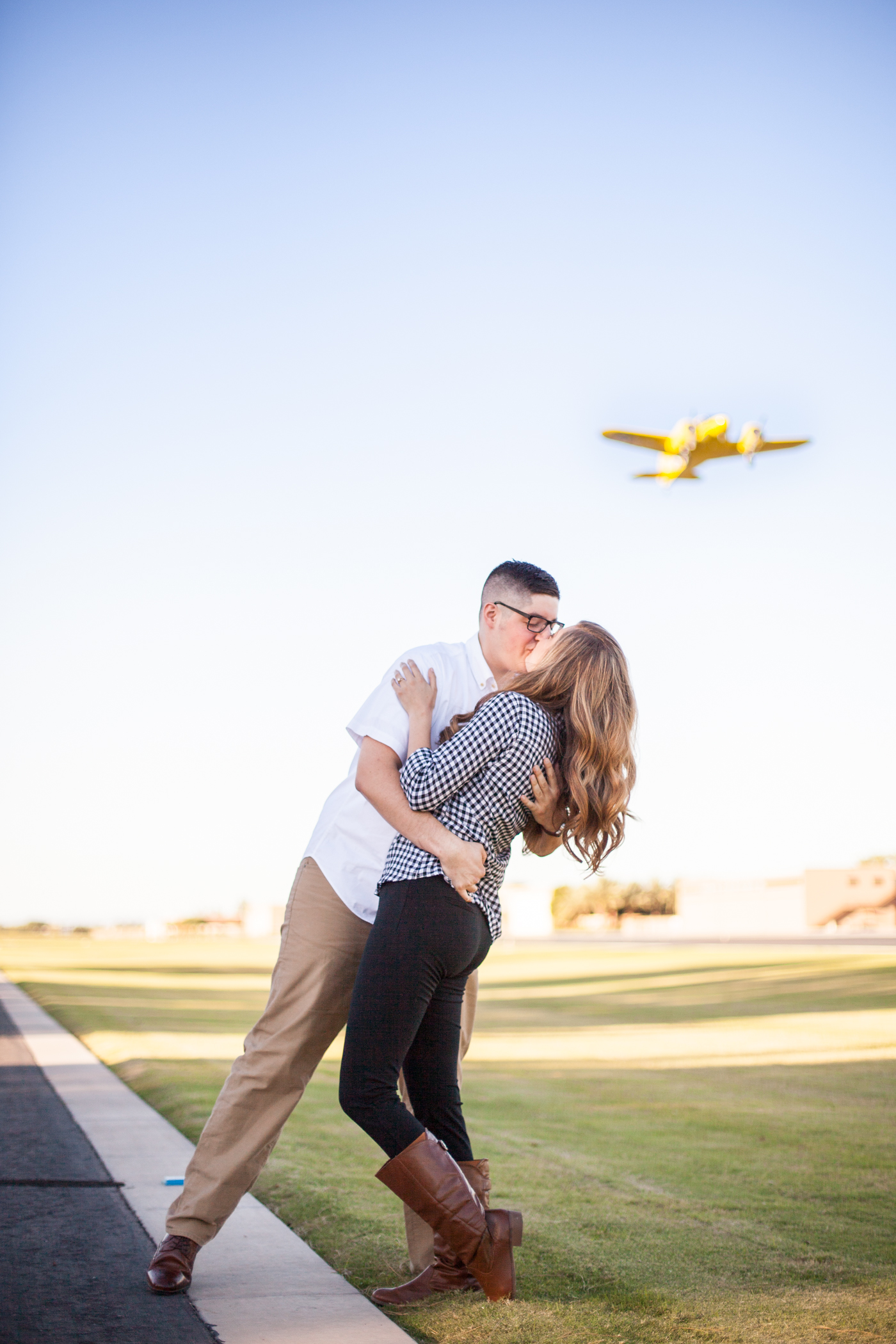 engaged couple kissing at a private airport with a plane flying above them