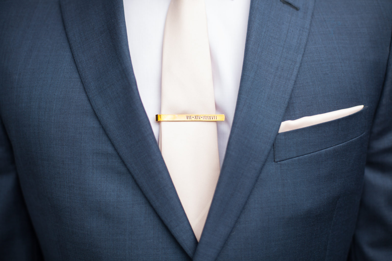 groom in navy blue suit with pink tie pocket square and personalized tie bar with wedding date engraved with roman numerals